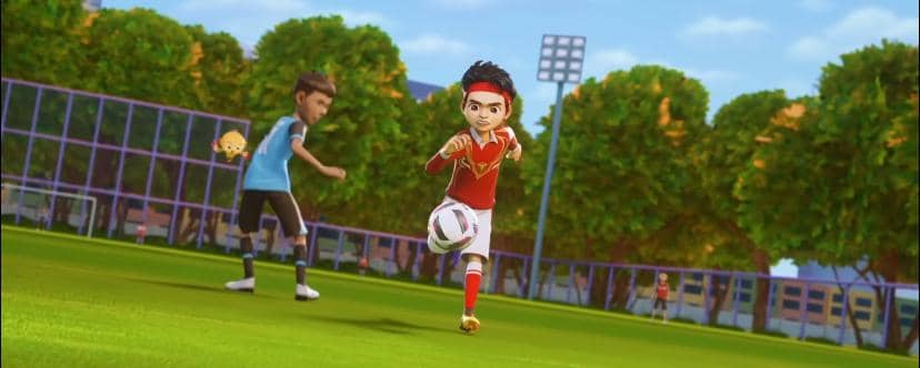 Karakter animasi mengenakan seragam merah berlari mengejar bola di lapangan sepak bola dengan latar pepohonan dan gawang.