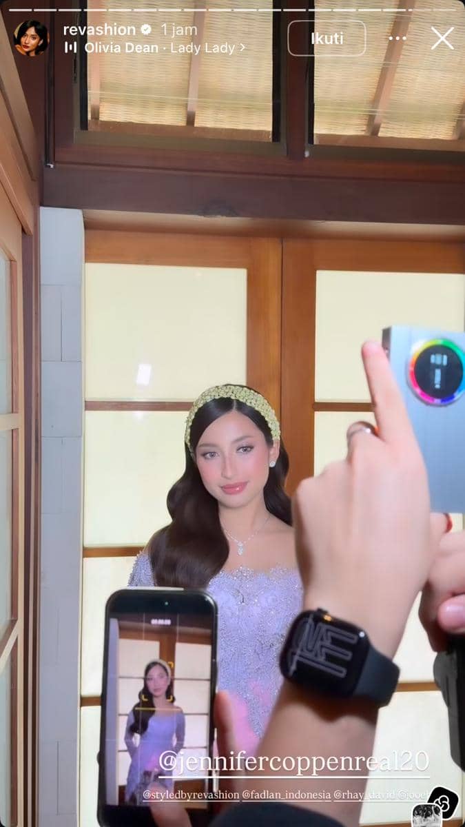 Seorang perempuan mengenakan kebaya ungu muda dengan hiasan kepala berwarna emas sedang berpose di depan kamera menjelang acara siraman.