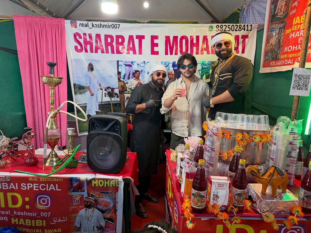 Tiga pria berdiri di stan minuman Sharbat E Mohabbat dengan dekorasi warna-warni, botol sirup, dan gelas plastik di atas meja.