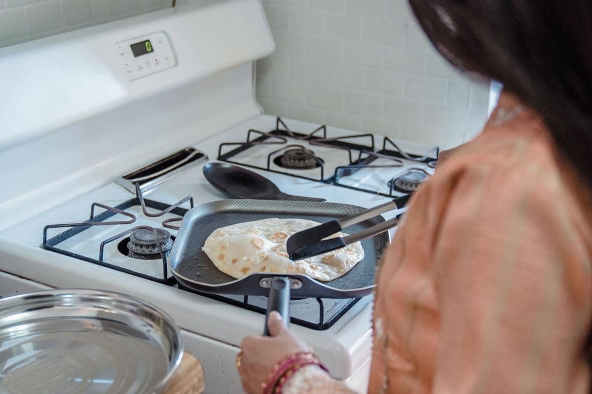 Seseorang sedang memanggang roti canai di atas wajan datar di kompor gas menggunakan penjepit logam di dapur rumah.