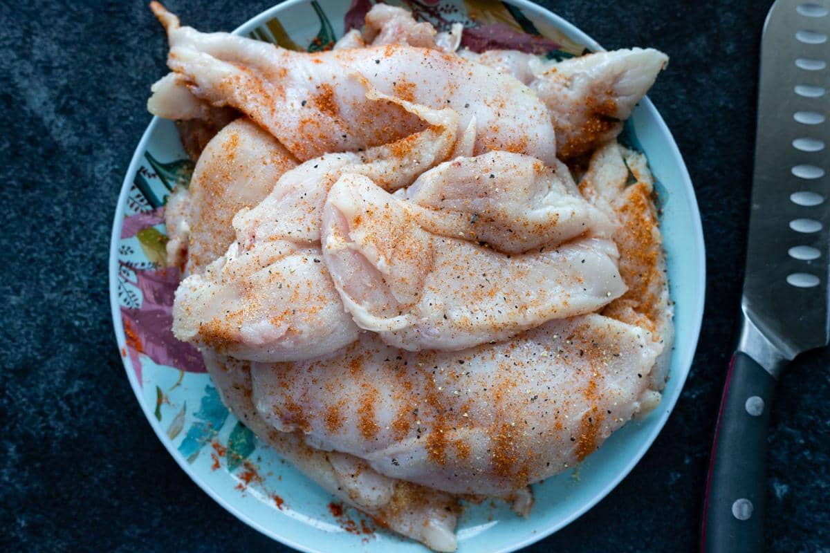 Potongan kulit ayam mentah yang telah dibumbui dengan rempah dan lada di atas piring, siap untuk dimasak.