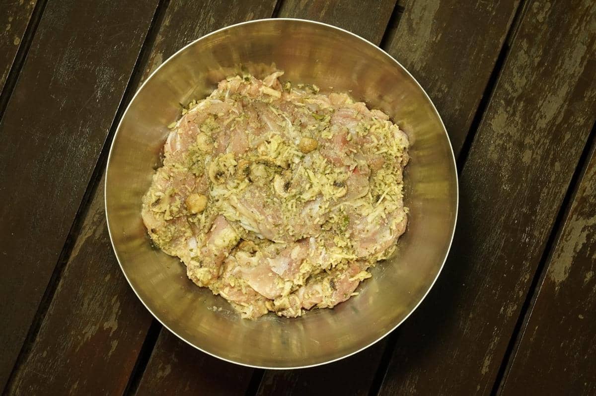 Kulit ayam yang sedang dimarinasi dengan bumbu rempah dalam mangkuk logam di atas meja kayu berwarna gelap.