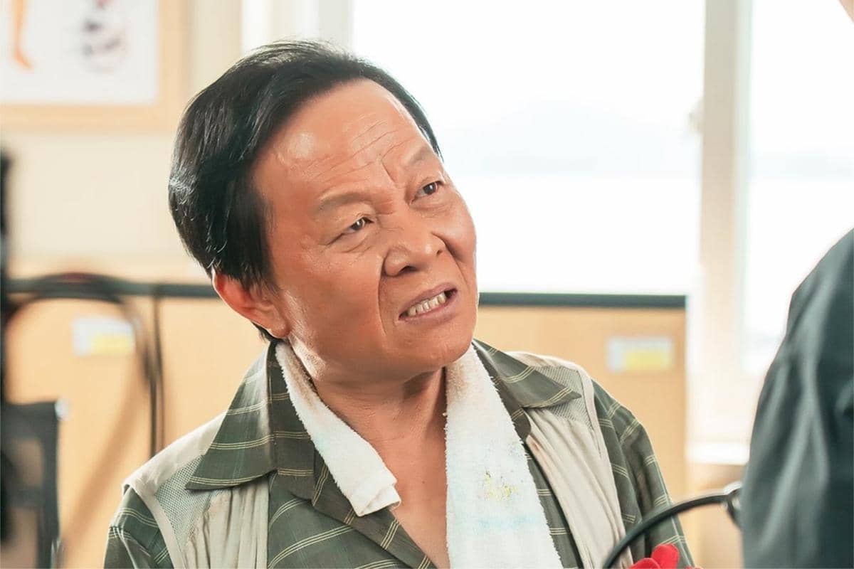 Seorang pria paruh baya mengenakan kemeja kotak-kotak dan handuk di lehernya sedang berbicara di dalam ruangan terang.