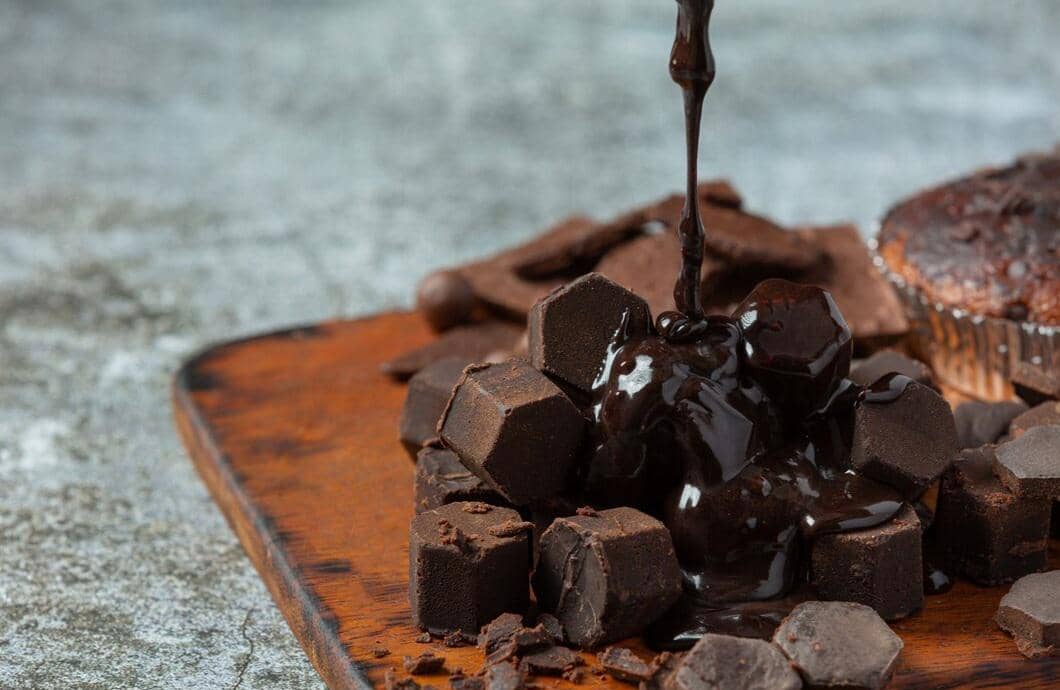 Potongan cokelat masak hitam disiram lelehan cokelat kental di atas papan kayu dengan latar abu-abu.
