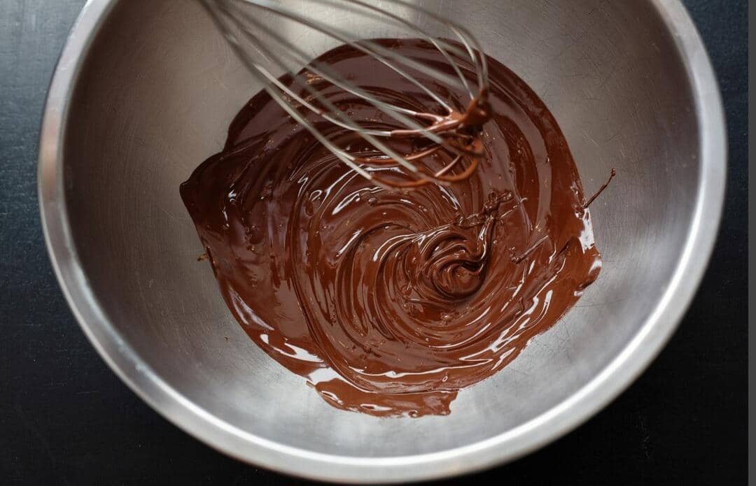 Cokelat leleh bertekstur lembut di dalam mangkuk logam dengan alat pengocok kawat di atas permukaannya.