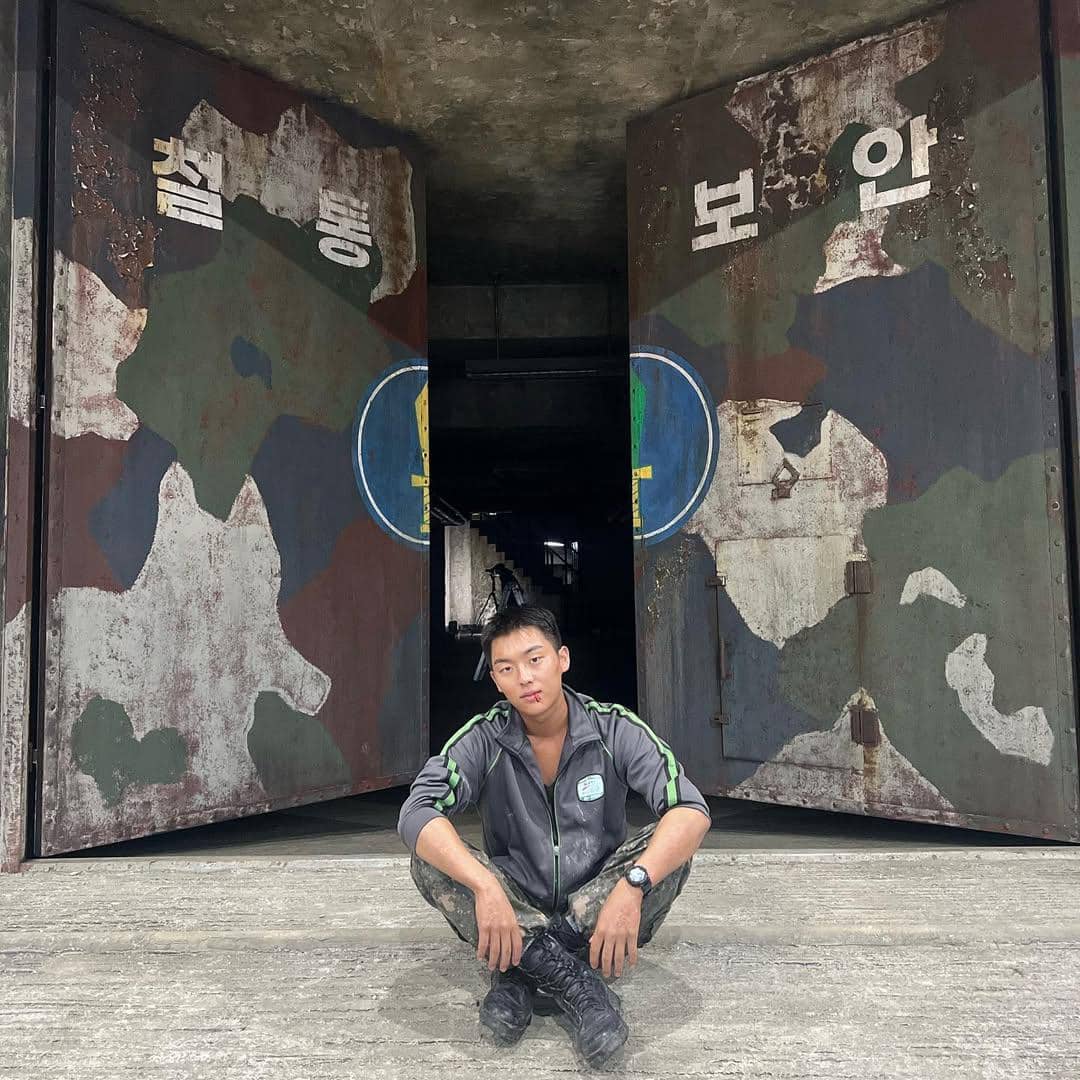 Seorang pria duduk di depan pintu besar bercorak kamuflase dengan tulisan Korea, mengenakan pakaian abu-abu dan sepatu hitam.