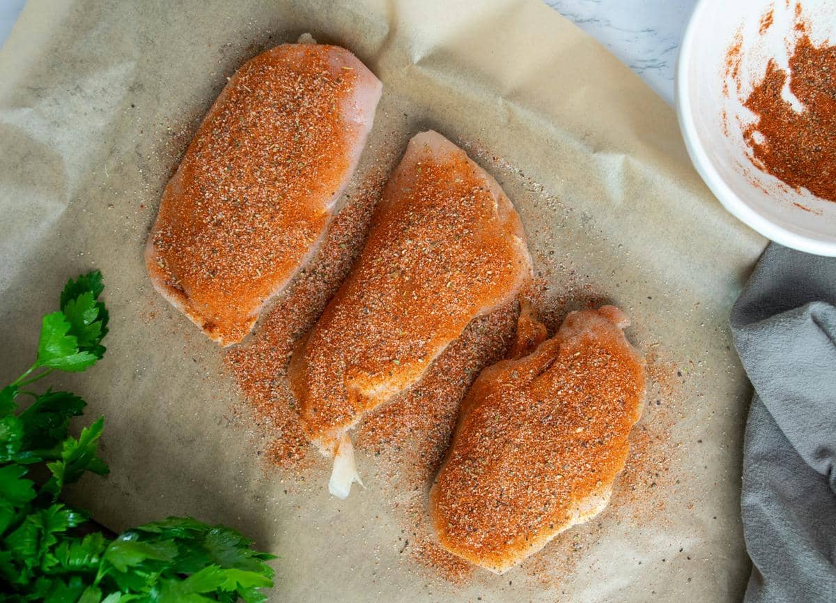 Tiga potong dada ayam mentah dilapisi bumbu marinasi berwarna merah kecokelatan di atas kertas roti dengan daun peterseli di sampingnya.