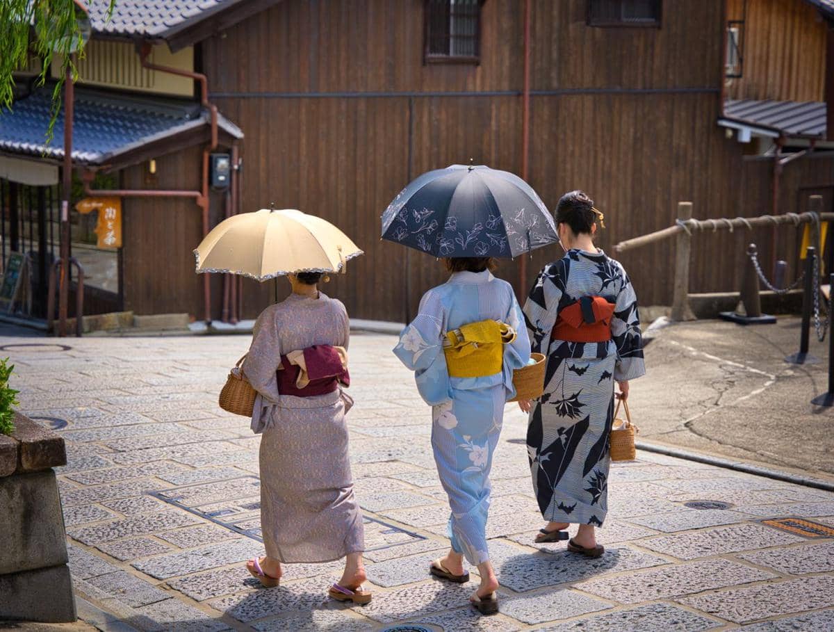 Tiga wanita mengenakan kimono tradisional Jepang berjalan di jalan berbatu sambil membawa payung di kawasan berarsitektur kayu.