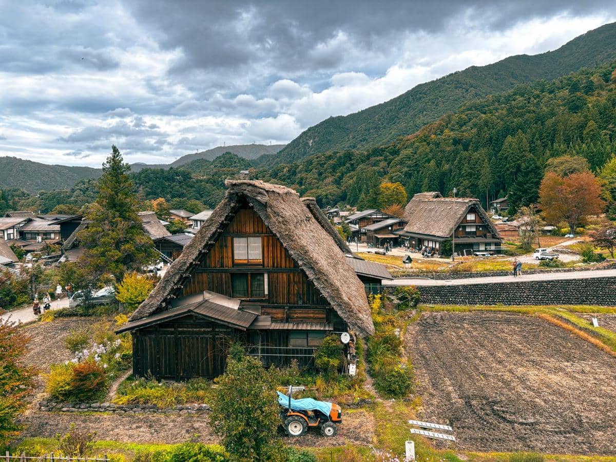 Rumah tradisional beratap jerami di pedesaan Jepang dengan latar pegunungan hijau dan sawah di bawah langit berawan.