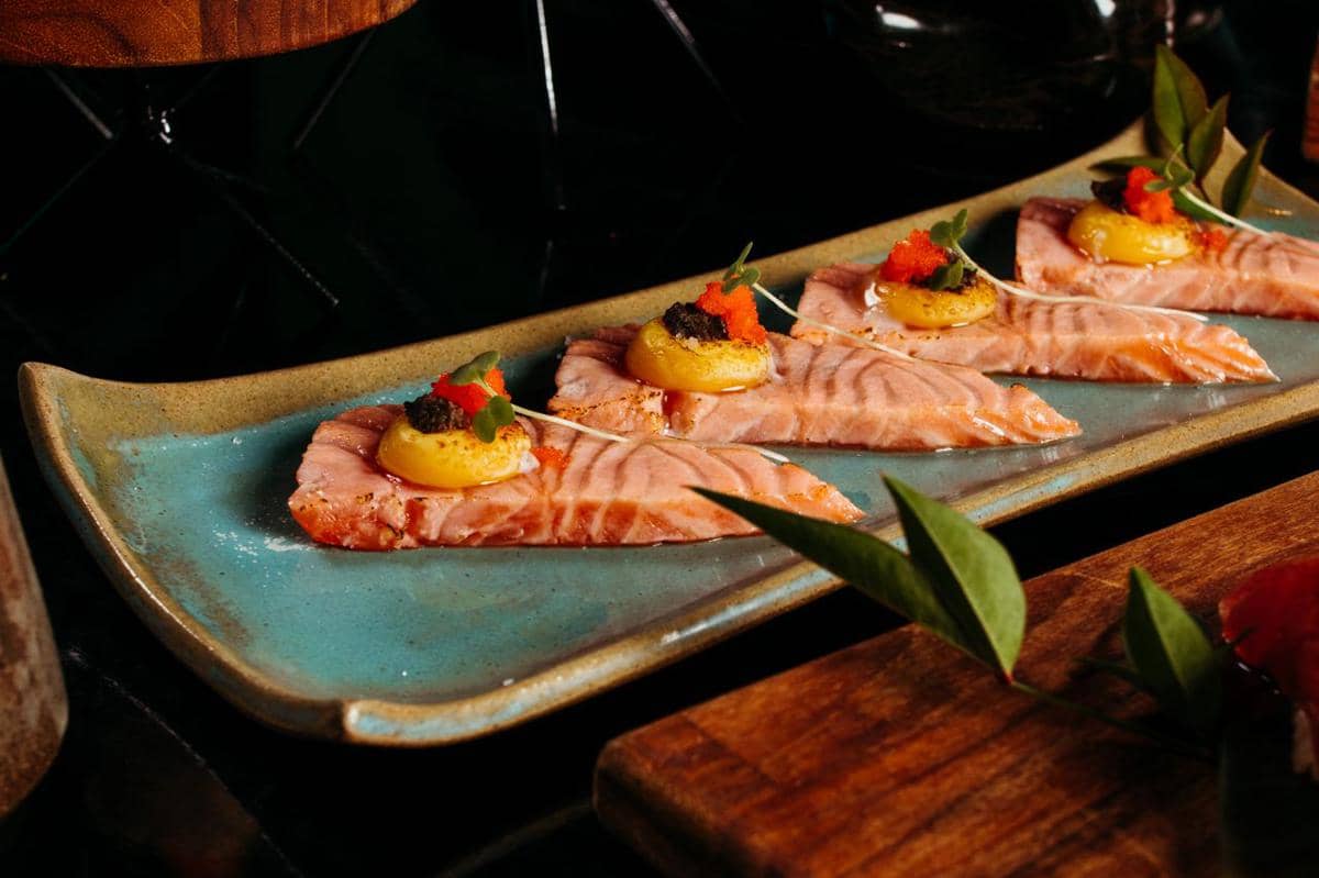 Potongan sashimi salmon disajikan di piring keramik biru dengan topping saus kuning, kaviar, dan daun kecil sebagai hiasan.