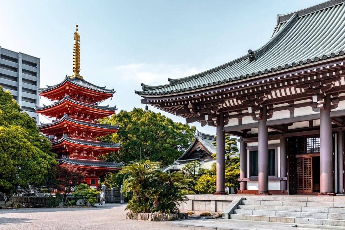 Bangunan kuil tradisional Jepang dengan pagoda merah bertingkat di antara pepohonan hijau pada siang hari yang cerah.