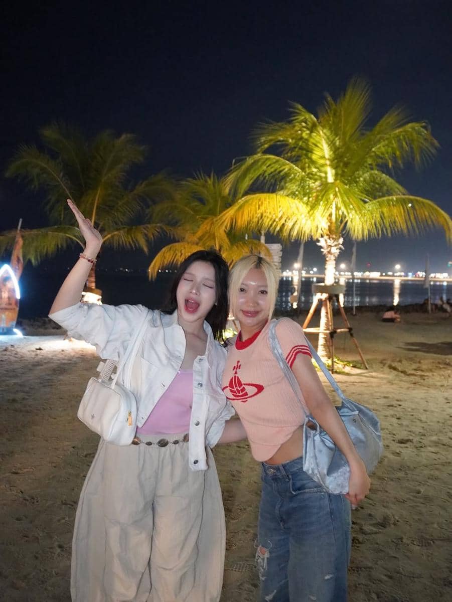 Dua perempuan berpose ceria di pantai pada malam hari dengan latar pohon kelapa dan lampu kota yang berkilau di kejauhan.