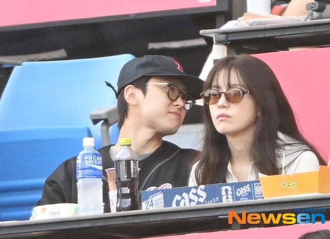 Dua orang duduk di tribun stadion baseball, mengenakan kacamata hitam dan pakaian kasual sambil menikmati suasana pertandingan.