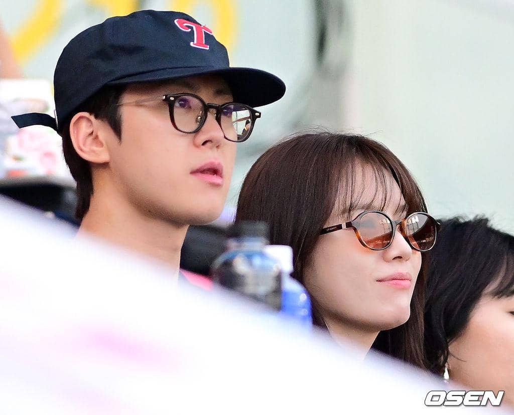 Dua orang mengenakan kacamata duduk berdampingan di tribun stadion baseball, menikmati pertandingan dengan suasana santai.