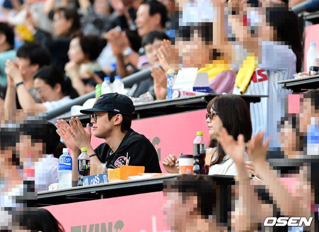 Dua orang duduk di tribun stadion baseball, mengenakan kacamata hitam dan pakaian kasual sambil menonton pertandingan bersama penonton lain.