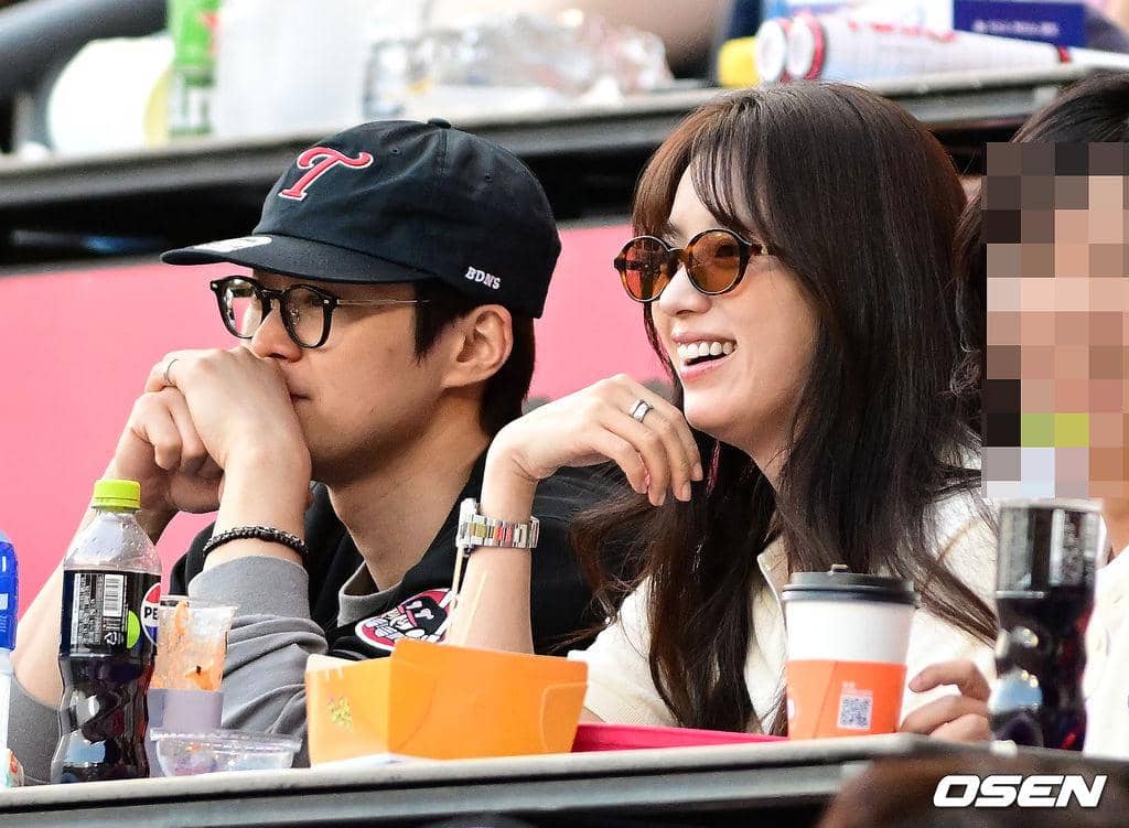 Dua orang duduk berdampingan di tribun stadion baseball, mengenakan kacamata hitam dan terlihat menikmati pertandingan dengan santai.