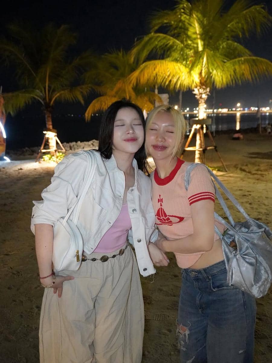 Dua perempuan berpose sambil tersenyum di pantai pada malam hari dengan latar pohon kelapa dan lampu kota di kejauhan.