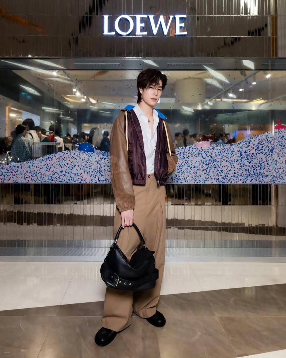 Seorang pria bergaya kasual elegan mengenakan jaket cokelat dan celana longgar sambil membawa tas hitam di depan toko Loewe.