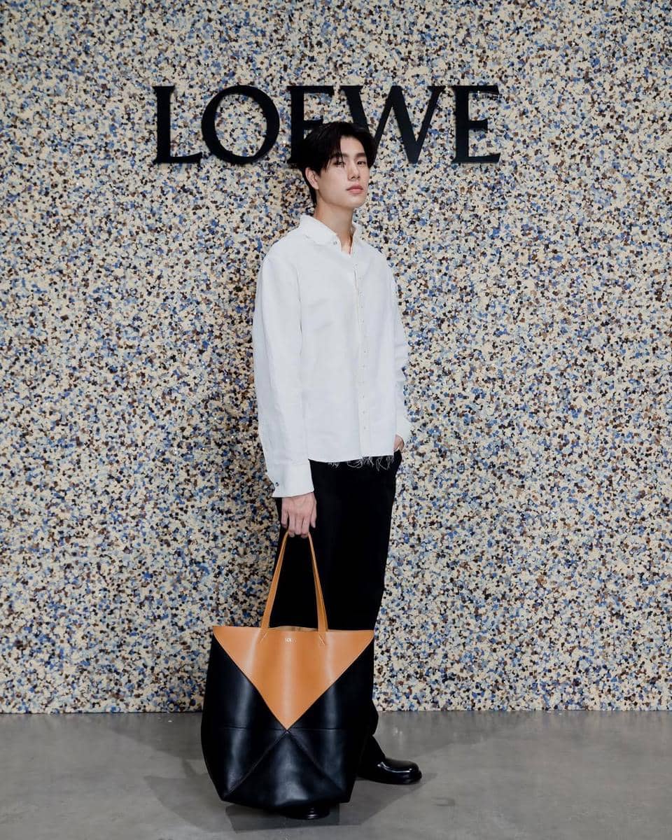Seorang pria mengenakan kemeja putih dan celana hitam sambil memegang tas besar berwarna hitam dan cokelat di depan dinding bermotif dengan logo Loewe.