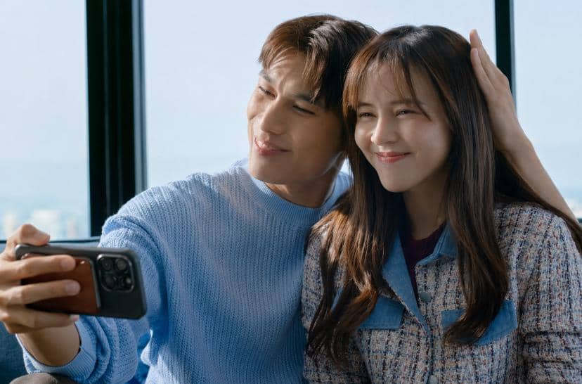 Sepasang karakter dalam drama My Wife's Gunslinger God tersenyum sambil berfoto selfie di dekat jendela besar dengan latar pemandangan luar.