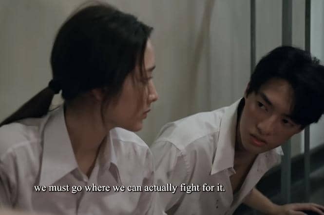 Dua siswa berseragam putih duduk di lantai dekat pagar besi, saling menatap serius dalam adegan drama When Light Fades.