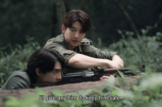 Dua pria berpakaian militer bersembunyi di balik semak di hutan, salah satunya memegang senapan sambil berbicara serius.