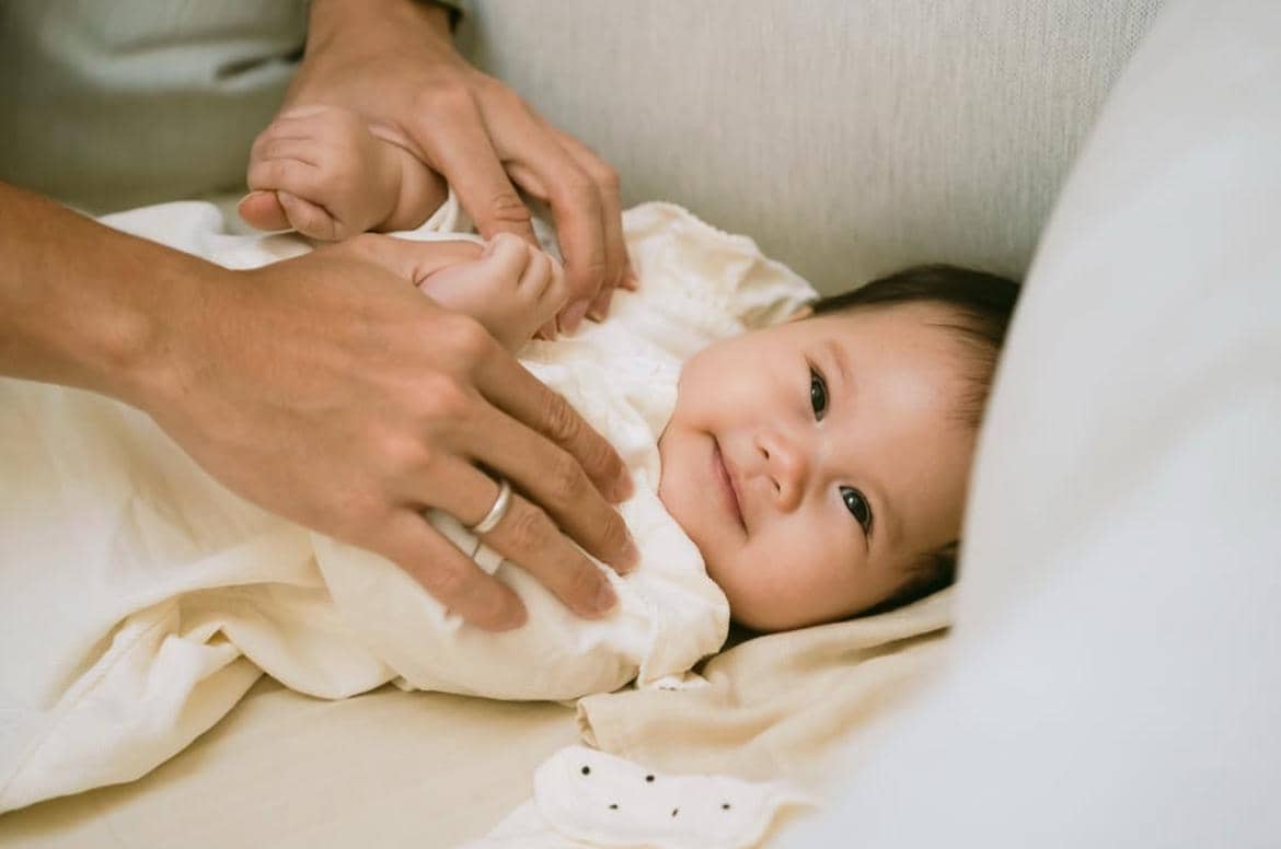 Bayi tersenyum manis sambil berbaring di tempat tidur dengan pakaian berwarna krem dan tangan orang dewasa menyentuhnya lembut.