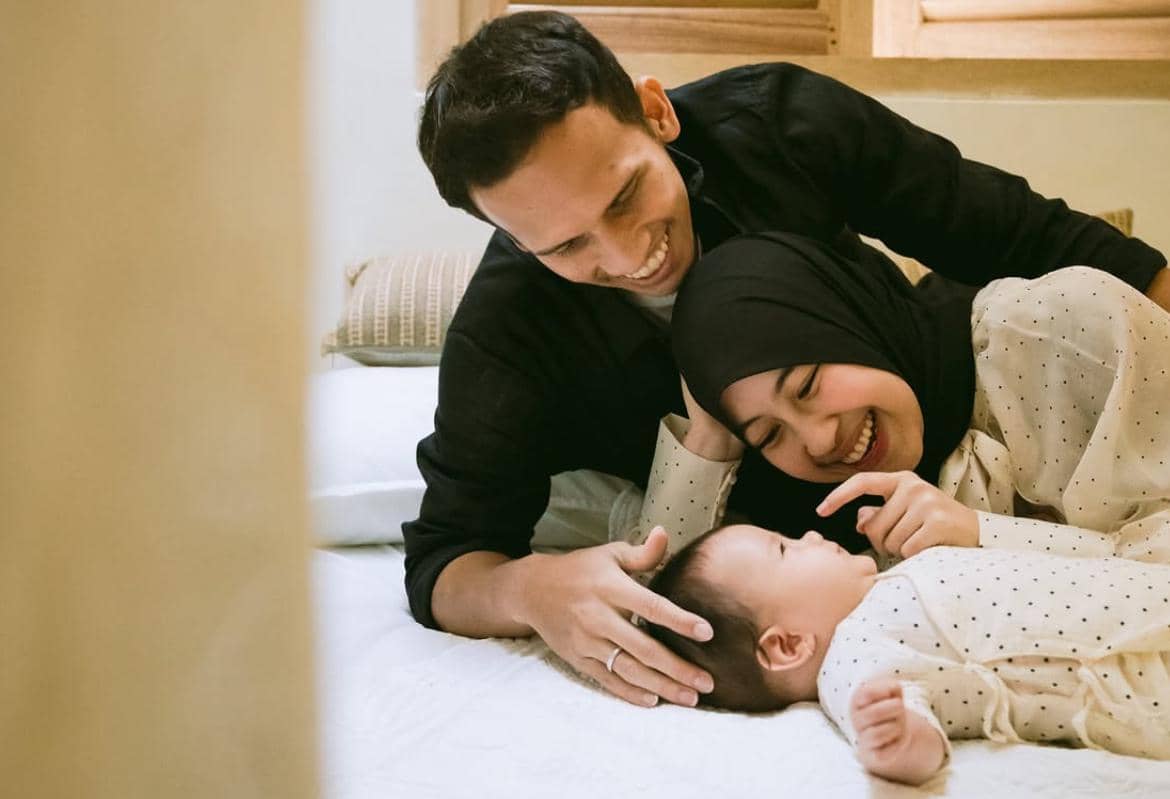 Sepasang orang dewasa tersenyum bahagia sambil berbaring di tempat tidur bersama bayi yang mengenakan pakaian putih bermotif polkadot.