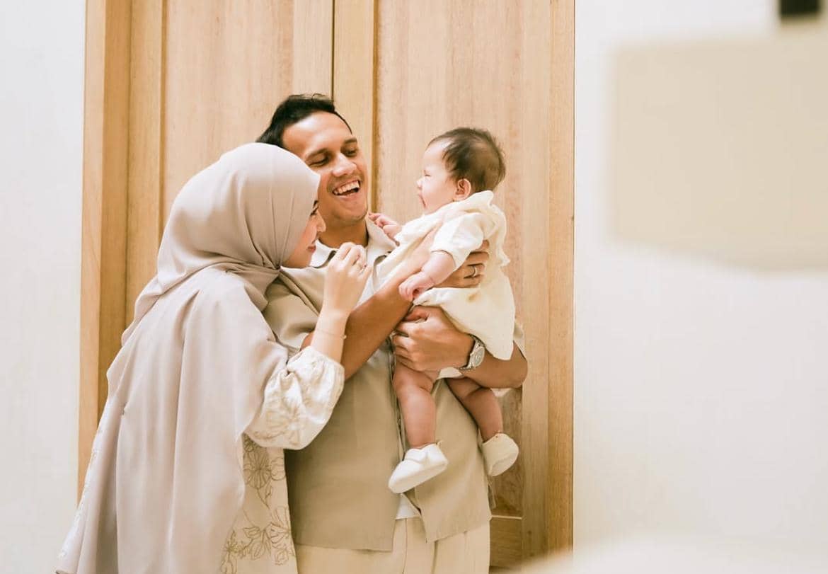 Seorang pria dan wanita tersenyum bahagia sambil menggendong bayi mereka di dalam ruangan dengan latar pintu kayu.