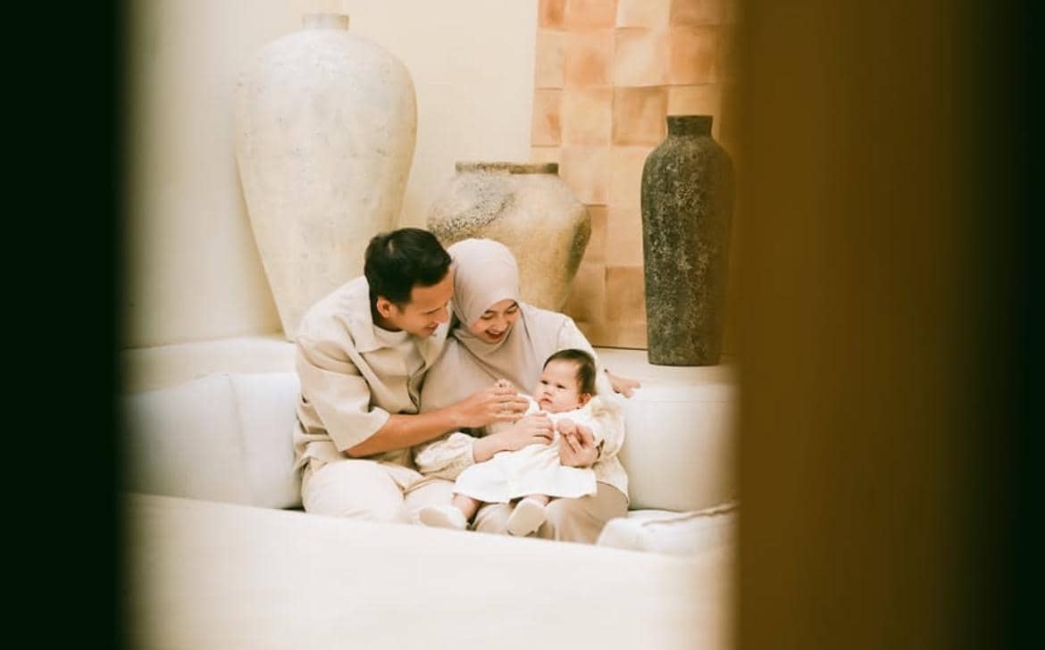 Sepasang orang dewasa duduk di sofa berwarna terang sambil tersenyum dan menggendong bayi di ruangan dengan dekorasi vas besar.
