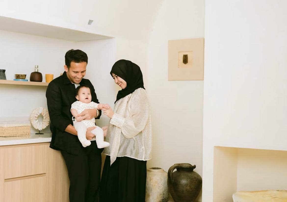 Sepasang orang dewasa tersenyum sambil menggendong bayi di ruangan bergaya minimalis dengan dekorasi vas dan rak kayu.