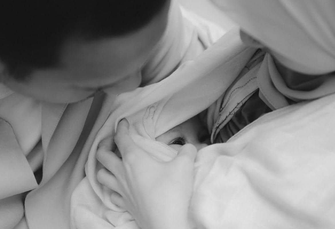 Seorang bayi yang dibungkus selimut tampak dipeluk hangat oleh dua orang dewasa dalam foto hitam putih penuh kehangatan.