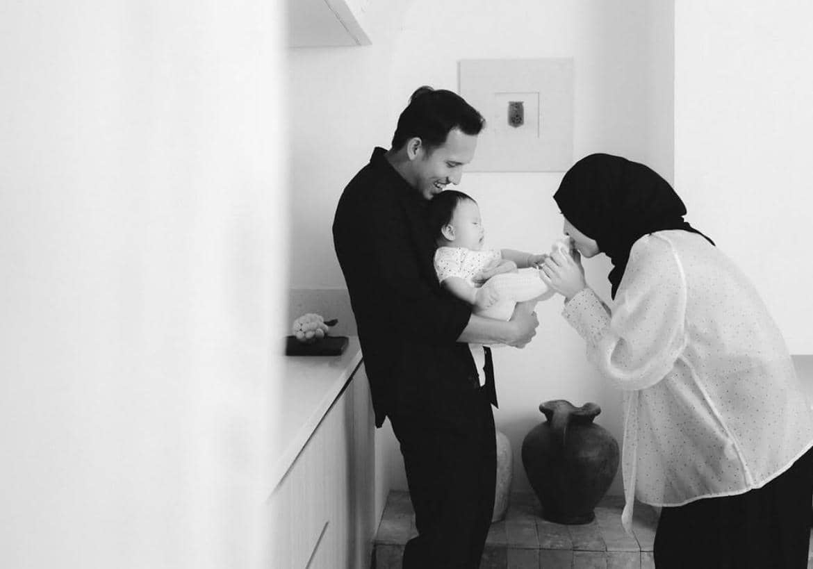 Seorang pria menggendong bayi sambil tersenyum, sementara seorang wanita berhijab mencium tangan bayi di ruangan bernuansa putih.