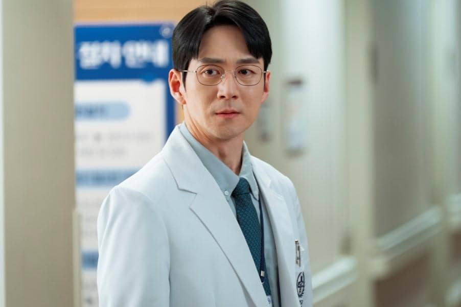Seorang pria mengenakan jas dokter putih dan dasi biru berdiri di koridor rumah sakit dalam cuplikan drama Reborn Rookie.