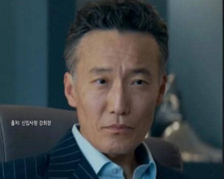 Seorang pria paruh baya mengenakan setelan jas bergaris duduk di kursi, menatap serius dalam cuplikan drama Reborn Rookie.