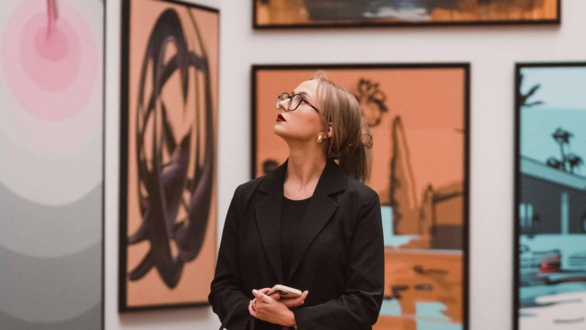Seorang perempuan berbusana hitam memegang ponsel sambil mengamati lukisan di dinding galeri seni atau museum modern.