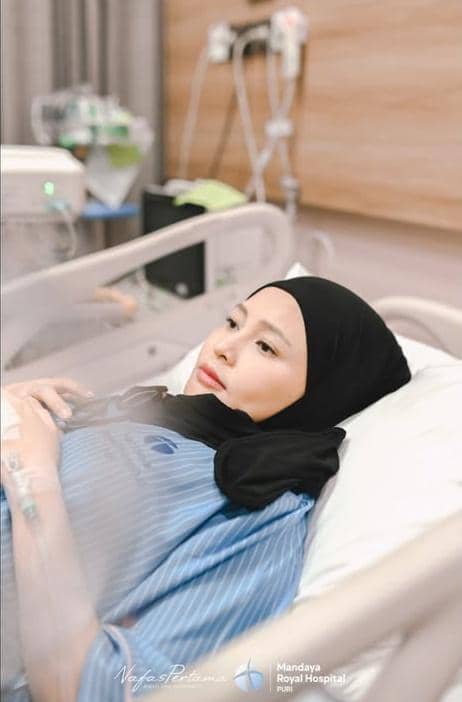 Seorang perempuan berhijab berbaring di ranjang rumah sakit dengan pakaian pasien, tampak tenang setelah melahirkan.