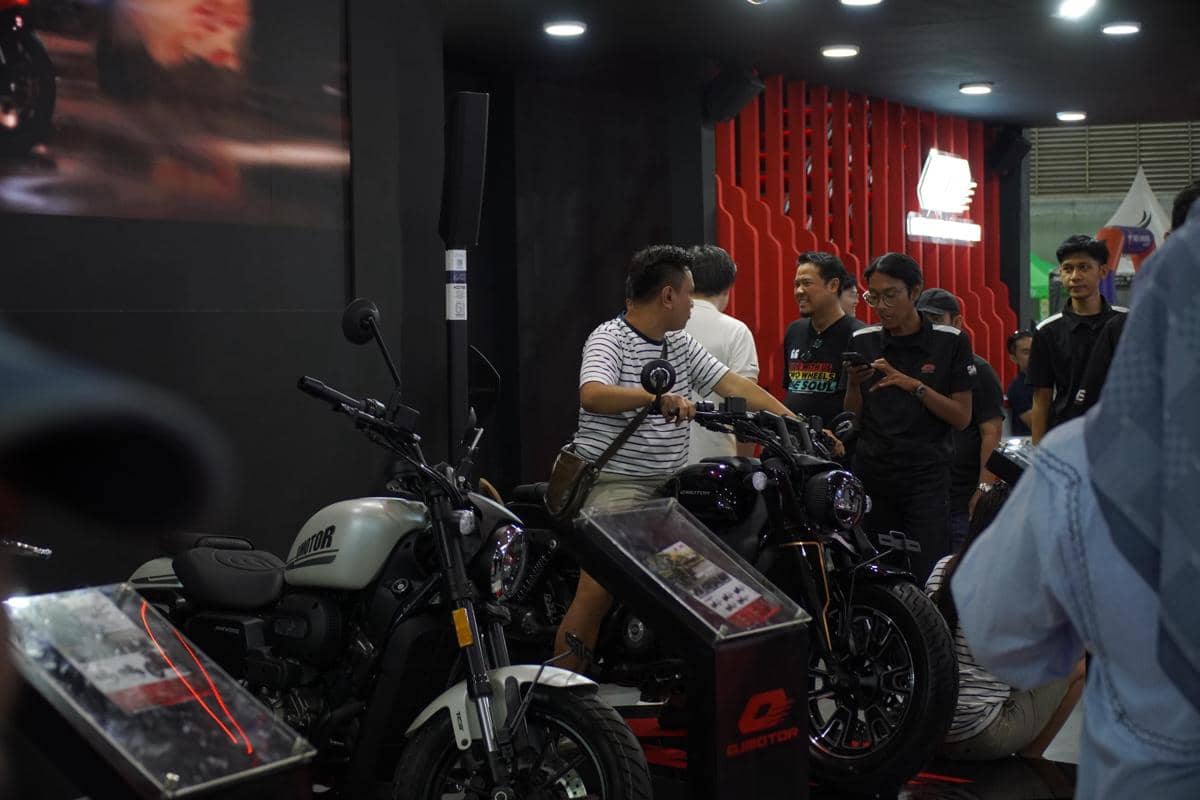 Pengunjung mencoba motor QJMotor di stan pameran dengan beberapa orang berbincang di area promosi dalam ruangan terang.