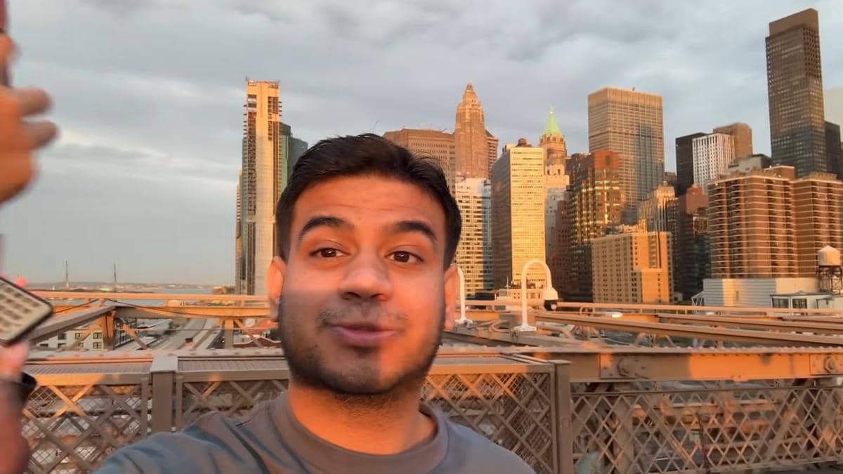 Seorang pria berpose di jembatan dengan latar gedung pencakar langit di Amerika Serikat saat matahari terbenam.