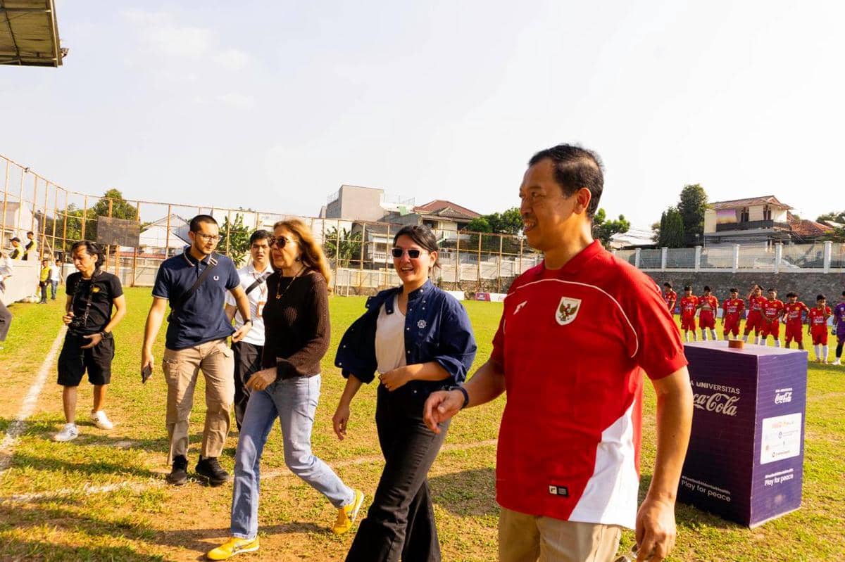 Beberapa orang berjalan di lapangan sepak bola saat acara Liga Universitas 2026 dengan tim berpakaian merah berbaris di belakang mereka.