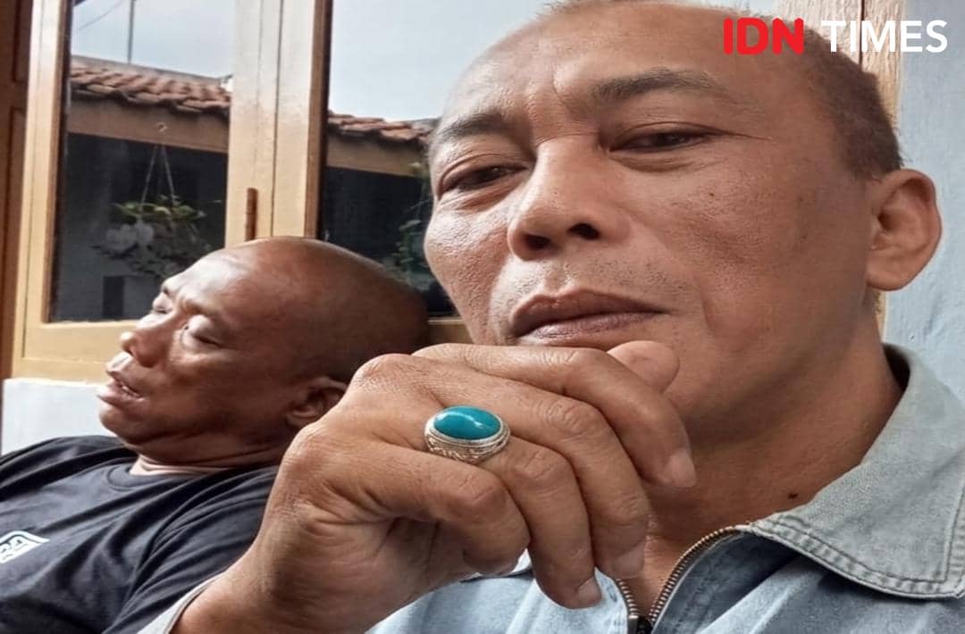 Seorang pria mengenakan cincin batu bacan berwarna biru di jarinya, duduk bersama seorang pria lain di dekat jendela kayu.
