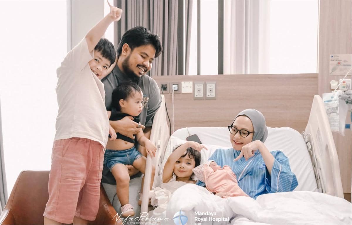 Seorang ibu di ranjang rumah sakit tersenyum sambil menggendong bayi baru lahir, didampingi suami dan tiga anak mereka yang tampak bahagia.