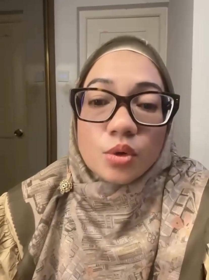 Seorang perempuan berhijab bermotif cokelat mengenakan kacamata besar berbingkai hitam sedang berbicara di dalam ruangan.
