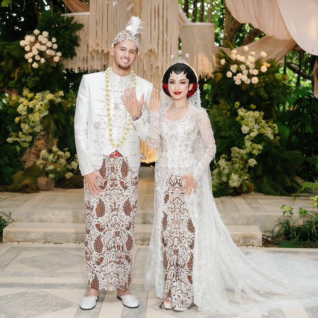 Pasangan pengantin mengenakan busana adat Jawa berwarna putih dan batik cokelat berpose di pelaminan dengan latar dekorasi bunga.
