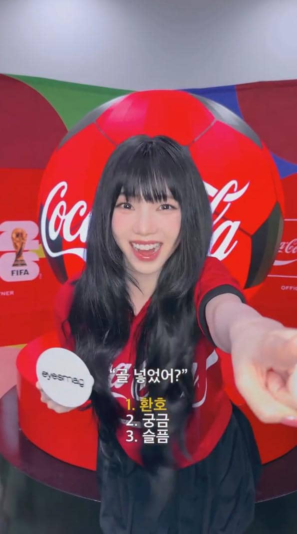 Seorang perempuan berambut panjang mengenakan jersey merah hitam berpose ceria di depan bola raksasa bertema Coca-Cola dan logo FIFA.