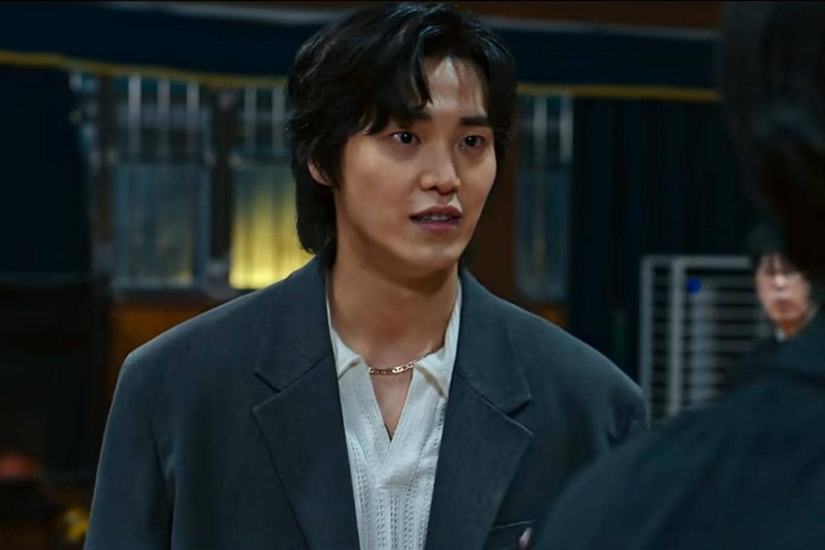 Seorang pria berambut hitam sedang berbicara di dalam ruangan dengan pencahayaan hangat dalam adegan drama Korea Teach You a Lesson.