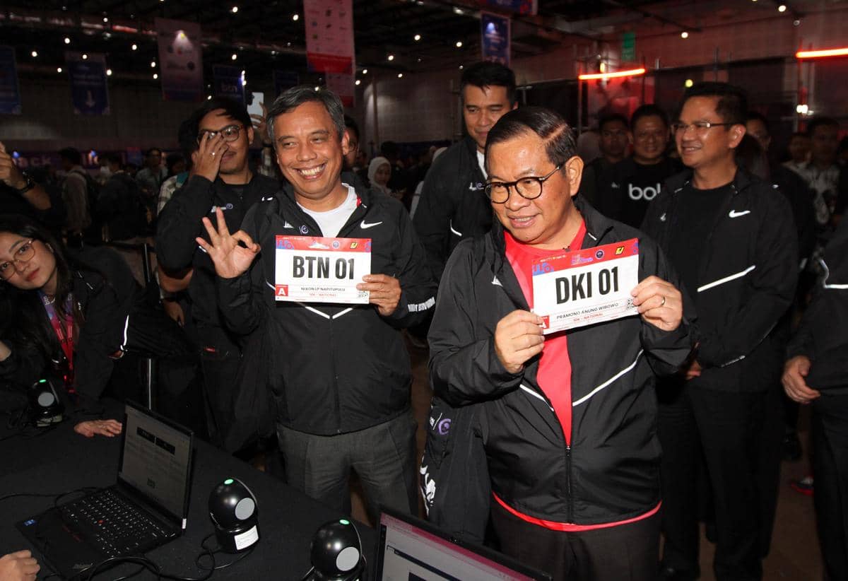 Seorang pejabat mengenakan jaket hitam memegang nomor peserta DKI 01 bersama peserta lain memegang BTN 01 di acara persiapan BTN JAKIM 2026.