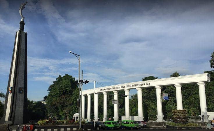 Tugu Kujang dan gerbang dengan deretan pilar putih di Kota Bogor pada siang hari dengan langit cerah dan kendaraan melintas di jalan.