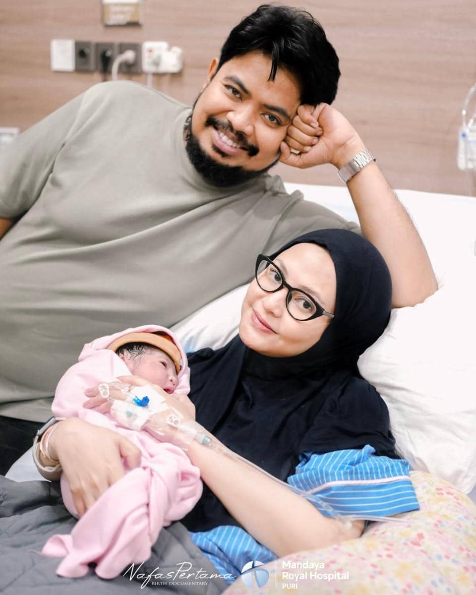 Seorang pria tersenyum di samping perempuan berhijab yang sedang menggendong bayi baru lahir di ruang rumah sakit.