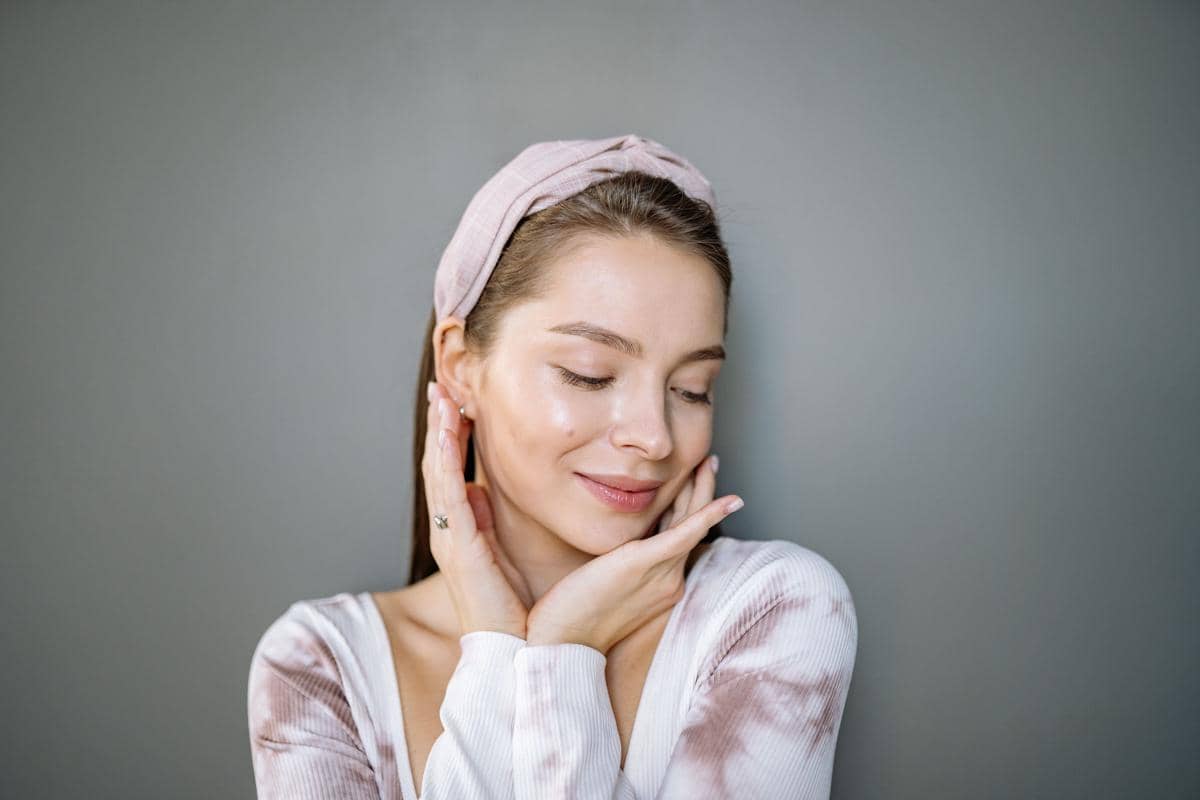 Seorang wanita dengan tampilan no makeup makeup, mengenakan ikat kepala berwarna lembut dan pakaian bermotif tie dye, tersenyum lembut.